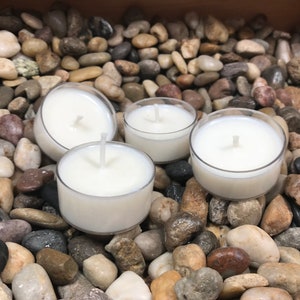 May include: Four white unscented tea light candles on a bed of small gray, brown, and white stones.