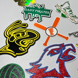 May include: Assortment of colorful iron-on patches with various designs, including a skull, a volleyball, a bird, a baseball bat, and a text that says "Lady Pirates".