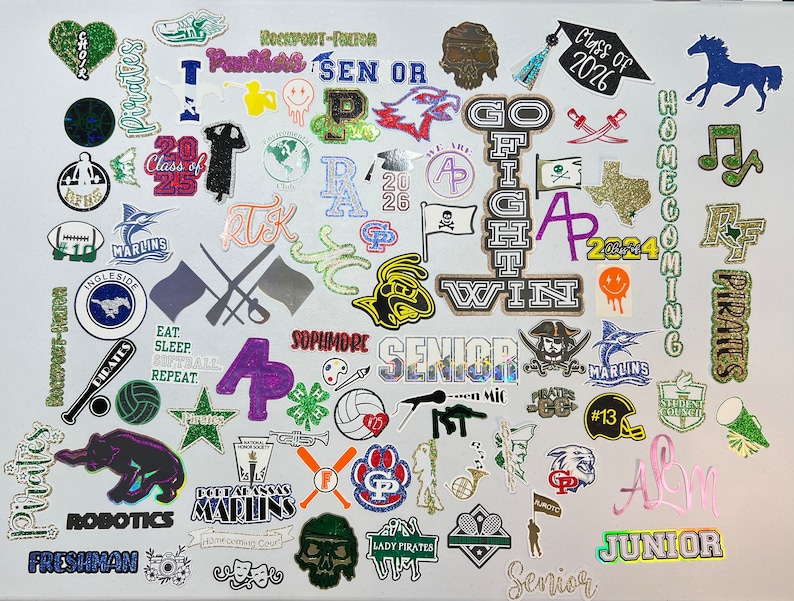 May include: A collection of colorful stickers featuring various school-related themes, including sports, clubs, and graduation years. The stickers are arranged in a random pattern on a white background. Some of the stickers have glitter and text, such as "Pirates", "Marlins", "Class of 2026", "Senior", "Freshman", "Junior", "Go Fight Win", "Homecoming", and "Robotics".