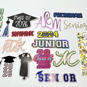 May include: A collection of colorful graduation themed iron-on patches. The patches feature various graduation year numbers, class of, senior, freshman, junior, sophomore, and homecoming.