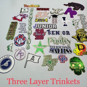 May include: A collection of 20 glitter-covered, three-layer trinkets with various school-themed designs. The designs include school mascots, class years, and school names like "Pirates", "Panthers", "Marlins", and "Sophmore".