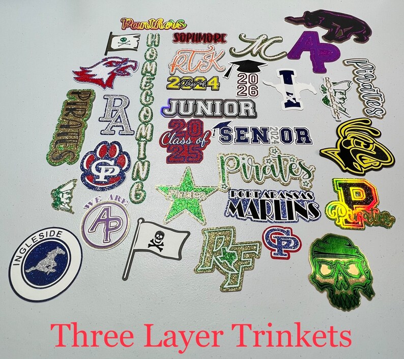 May include: A collection of 20 glitter-covered, three-layer trinkets with various school-themed designs. The designs include school mascots, class years, and school names like "Pirates", "Panthers", "Marlins", and "Sophmore".
