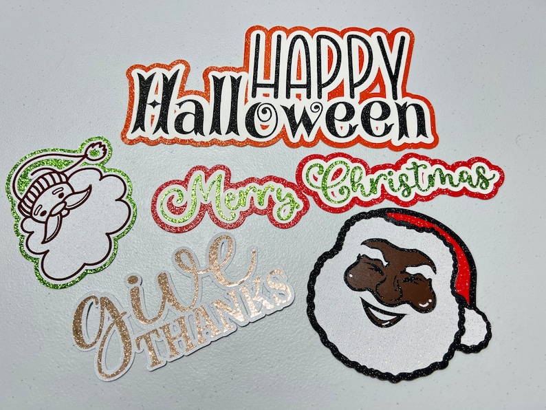 May include: Five glitter-covered paper cutouts with holiday greetings. The cutouts are in various colors, including red, green, gold, and black. The greetings are "Happy Halloween", "Merry Christmas", "Give Thanks", and a Santa Claus face.