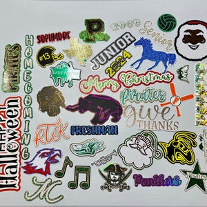 May include: A collection of colorful, glittery iron-on patches with various school-related themes, including "Homecoming", "Pirates", "Senior", "Junior", "Freshman", "Lady Pirates", "Give Thanks", "Merry Christmas", and "Halloween".