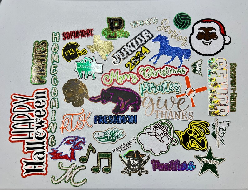 May include: A collection of colorful, glittery iron-on patches with various school-related themes, including "Homecoming", "Pirates", "Senior", "Junior", "Freshman", "Lady Pirates", "Give Thanks", "Merry Christmas", and "Halloween".
