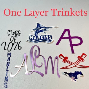 May include: A collection of one-layer trinkets, including a purple "A" with glitter, a purple "Alum" with glitter, a red crossed swords design, a blue and white marlin logo with the text "Marlins", a blue and white horse logo, and a black and white "Class of 2026" design with a blue glitter "Marlins" design.