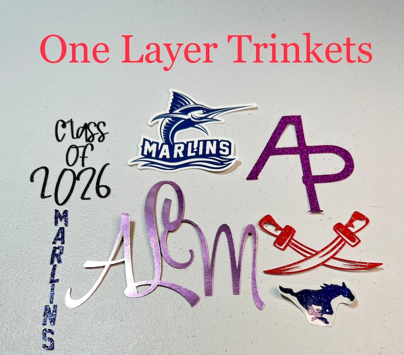May include: A collection of one-layer trinkets, including a purple "A" with glitter, a purple "Alum" with glitter, a red crossed swords design, a blue and white marlin logo with the text "Marlins", a blue and white horse logo, and a black and white "Class of 2026" design with a blue glitter "Marlins" design.