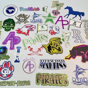 May include: A collection of colorful iron-on patches featuring various school mascots and logos, including pirates, panthers, and marlins. The patches are decorated with glitter and feature a variety of colors, including blue, green, red, gold, and purple.