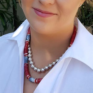 May include: A woman wearing a white collared shirt with a layered necklace. The necklace is made of red and blue beads and silver round beads.