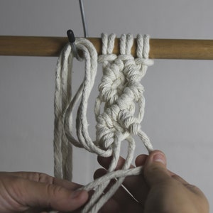 Pu&ograve; includere: Primo piano di un progetto di macram&egrave; bianco in corso. Il nodo viene fatto su un'asta di legno. Il nodo viene fatto con un cordoncino bianco.