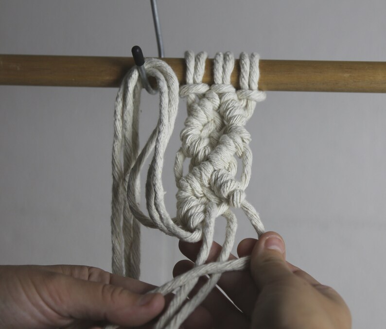 Pu&ograve; includere: Primo piano di un progetto di macram&egrave; bianco in corso. Il nodo viene fatto su un'asta di legno. Il nodo viene fatto con un cordoncino bianco.