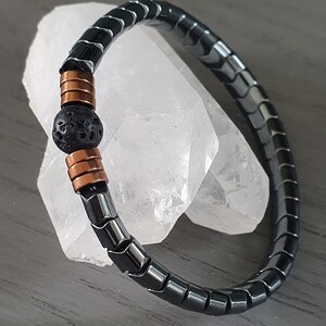 Może przedstawiać: Bransoletka z czarnego hematytu z czarną koralikiem lawy i miedzianymi rozdzielaczami. Bransoletka jest elastyczna i można ją dopasować do większości nadgarstków.