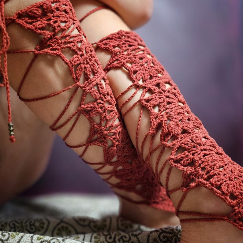 May include: Rust-coloured crochet leg wraps with intricate openwork patterns and delicate cord ties. The handmade wraps feature a floral design and are suitable for a bohemian or festival style. The wraps are designed to be worn on the legs.