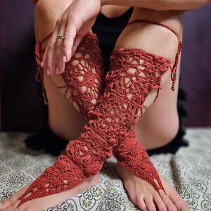 May include: Rust-coloured crochet leg wraps with a floral pattern. The wraps extend from the ankle to the thigh, with a toe loop and tie closure. The openwork design allows for breathability and a unique look. The wraps are handmade.