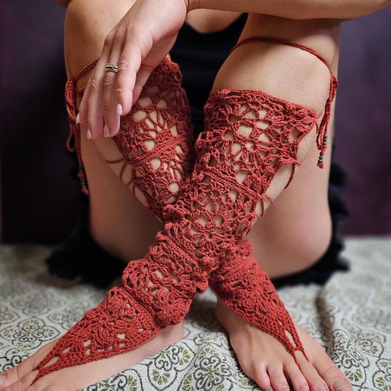 May include: Rust-coloured crochet leg wraps with a floral pattern. The wraps extend from the ankle to the thigh, with a toe loop and tie closure. The openwork design allows for breathability and a unique look. The wraps are handmade.