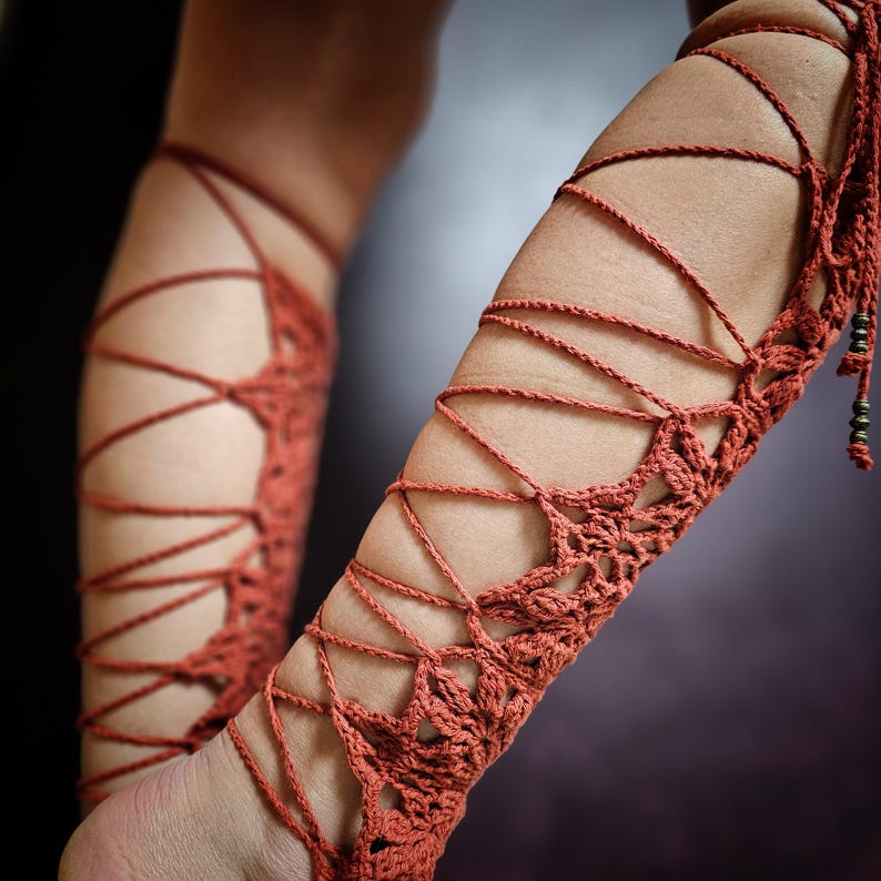 May include: Rust-coloured, handmade crochet arm cuffs. The cuffs feature a detailed lace-up design with intricate patterns and beaded accents. The crochet work is visible, showcasing the texture and craftsmanship. The cuffs are designed to be worn on the arm.