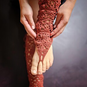 May include: Rust-coloured crochet foot jewellery with a floral design. The openwork pattern extends up the leg, creating a unique barefoot sandal look. The intricate details and warm colour make it a stylish accessory for beach or festival wear.