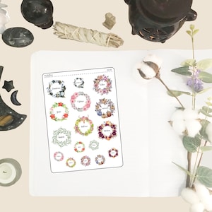 May include: A sheet of stickers featuring watercolor wreath designs with floral and botanical elements. Each wreath is labeled with a word, such as "Sagittarius" and "Libra". The stickers are in various colors, including pink, green, and purple.