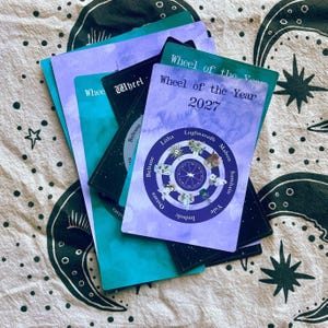 May include: A stack of rectangular cards featuring the text Wheel of the Year 2027. The top card is purple with a circular diagram showing seasonal labels like Litha, Mabon, and Yule, surrounded by small illustrations of flowers and nature symbols.