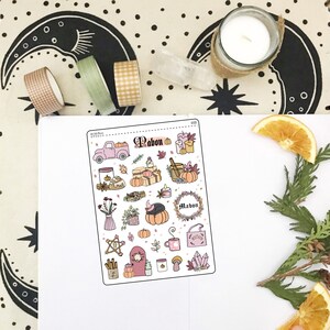May include: A sheet of pink and white stickers with a fall harvest theme. The stickers feature a truck, pumpkins, candles, a cauldron, a wreath, and other fall-themed imagery. The text "Mabon" is featured on the sheet.