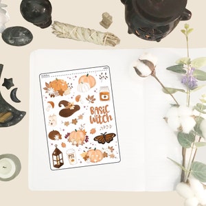 May include: A sheet of stickers with a fall and witch theme. The stickers feature pumpkins, leaves, a witch's hat, a butterfly, a lantern, a coffee cup, and a jar of honey. The text "Basic Witch" is written in brown lettering.