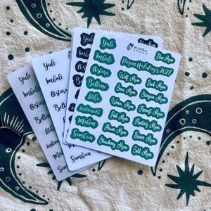May include: Four stacked sticker sheets featuring white backgrounds and green or black text. The top sheet displays the text Pagan Holidays 2027, Wise Moon Studio, and various holiday and moon names like Yule, Imbolc, Ostara, Beltane, and Blue Moon.