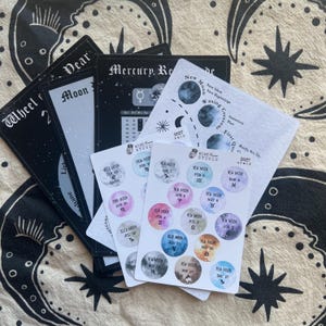 May include: A collection of printed paper sheets featuring 2027 lunar calendars and sticker sets. The items include black and white charts with text like Mercury Retrograde and Moon Phases, alongside two white sticker sheets with colorful circular moon icons.