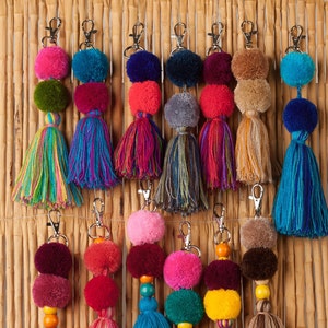 May include: A collection of thirteen colorful yarn keychains featuring various combinations of fuzzy pom-poms, wooden beads, and long hanging tassels attached to silver-toned metal lobster clasps. The text MexicanBeautyShop is visible at the bottom.