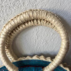 May include: Close-up of a woven straw bag handle. The handle is made of natural straw and is double-layered. The handle is attached to a colorful woven bag.