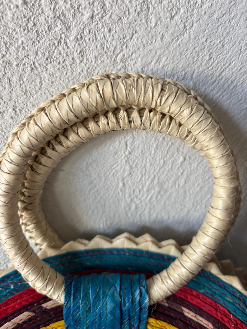 May include: Close-up of a woven straw bag handle. The handle is made of natural straw and is double-layered. The handle is attached to a colorful woven bag.