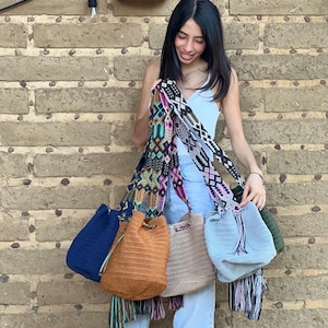 May include: Four crocheted bucket bags with colorful tassels. The bags are in shades of blue, brown, beige, and light blue. The bags are hanging from a person's arm.