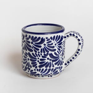 May include: White ceramic mug with a blue floral pattern. The mug has a rounded shape, a blue rim, and a handle decorated with blue dots. The floral design covers the body of the mug.
