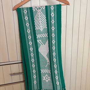 May include: A green and white woven scarf with a geometric pattern and fringe. The scarf is hanging on a wooden hanger. The design features white diamond and tree-like shapes on a green background.