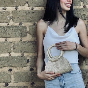 Op de afbeelding: Een vrouw houdt een klein, wit, geweven tasje vast met een ronde handgreep. De tas is gemaakt van natuurlijke materialen en heeft een gefranst randje. De vrouw draagt een wit tanktopje en een blauwe spijkerbroek.