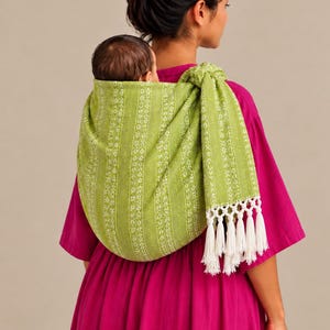 May include: A lime green baby carrier sling featuring a white floral and striped pattern with a knotted shoulder strap and long white fringe tassels at the end. The fabric has a textured appearance and is draped across the back.