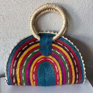 May include: A colorful half-circle woven straw bag with a large, round, woven handle. The bag is made of different colored woven strips, including red, yellow, green, blue, and purple.