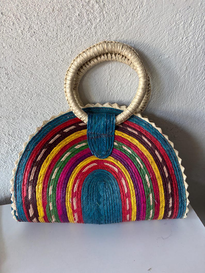 May include: A colorful half-circle woven straw bag with a large, round, woven handle. The bag is made of different colored woven strips, including red, yellow, green, blue, and purple.