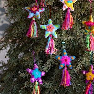 May include: Colorful star-shaped Christmas ornaments with tassels hanging from a green tree. The ornaments are made of felt and feature various colors, including blue, pink, yellow, and purple. Each star has a unique floral design in the center.