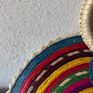 May include: A colorful woven straw plate with a circular design. The plate is made of multiple layers of straw, each layer a different color. The colors are blue, red, yellow, green, purple, and black.