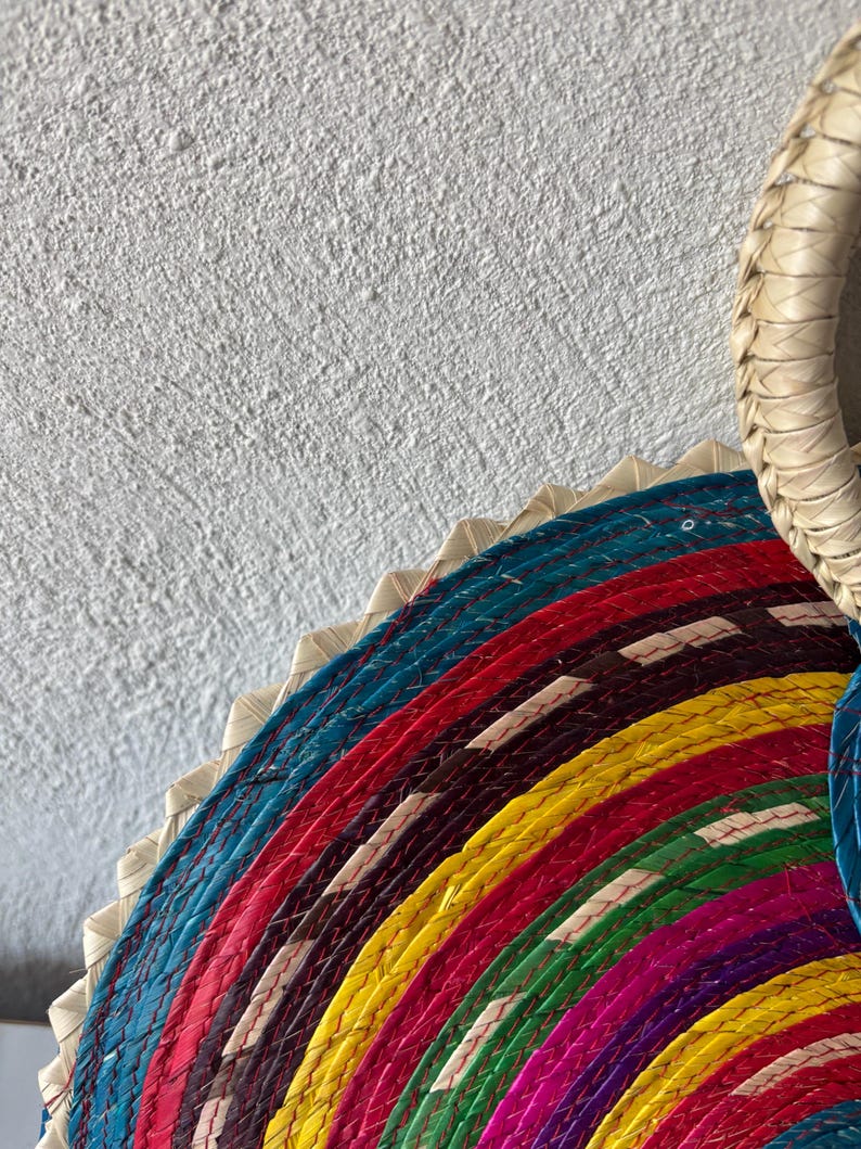 May include: A colorful woven straw plate with a circular design. The plate is made of multiple layers of straw, each layer a different color. The colors are blue, red, yellow, green, purple, and black.