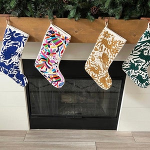 May include: Four Christmas stockings with embroidered designs hang above a fireplace. The stockings feature white fabric with colourful patterns, including blue animals, vibrant floral designs, gold birds, and green birds. The stockings are suspended from a wooden mantel.