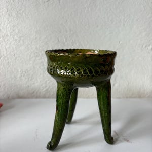 May include: A small, green ceramic pot with three legs. The pot has a decorative design around the top edge.