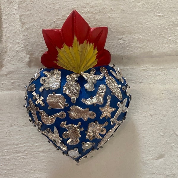 Puede incluir: Un adorno de cerámica azul en forma de corazón con una llama roja y amarilla en la parte superior. El corazón está decorado con dijes plateados, incluyendo estrellas, cruces y otros símbolos religiosos.