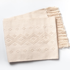 Puede incluir: Una alfombra tejida de color crema con un patrón geométrico y flecos en los bordes.