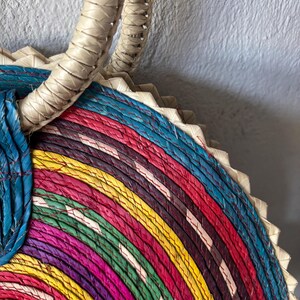 May include: A colorful woven straw bag with a thick, white handle. The bag is made of multiple layers of straw, each dyed a different color, creating a rainbow effect.