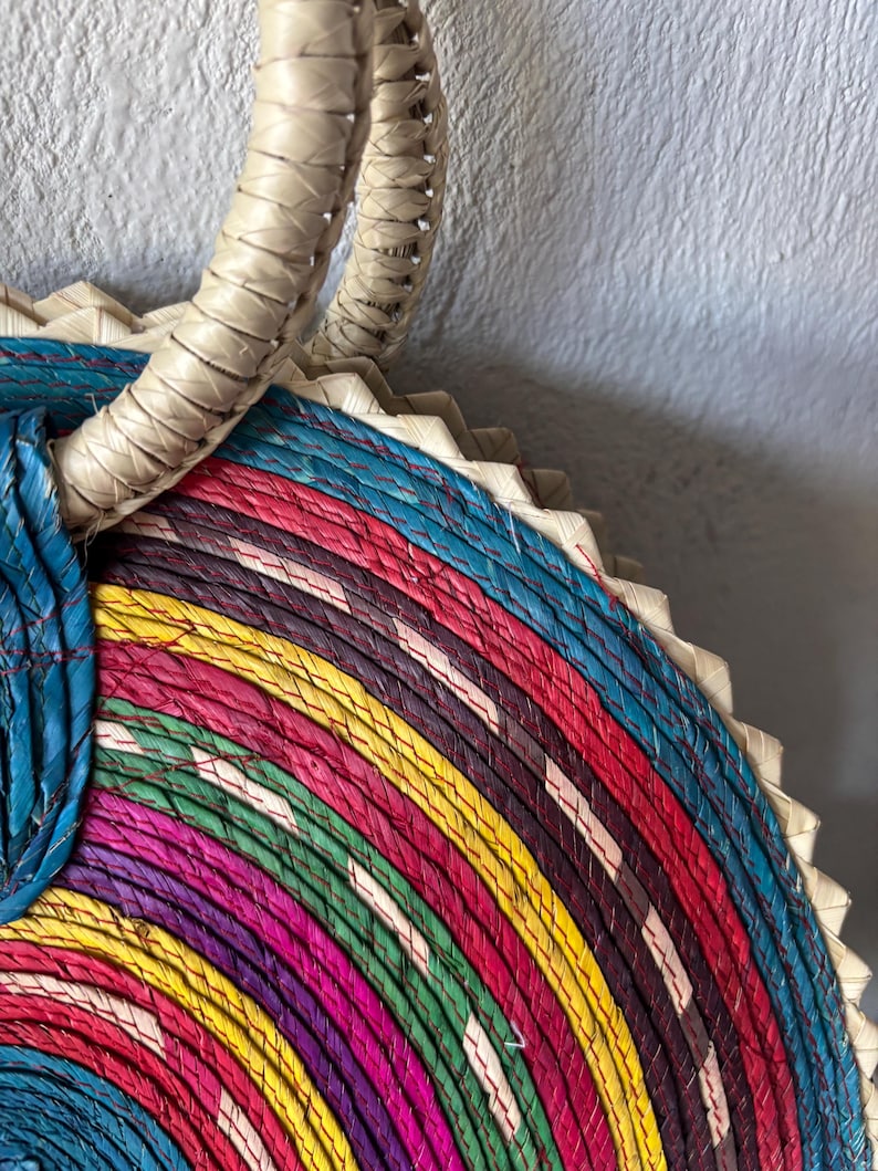 May include: A colorful woven straw bag with a thick, white handle. The bag is made of multiple layers of straw, each dyed a different color, creating a rainbow effect.