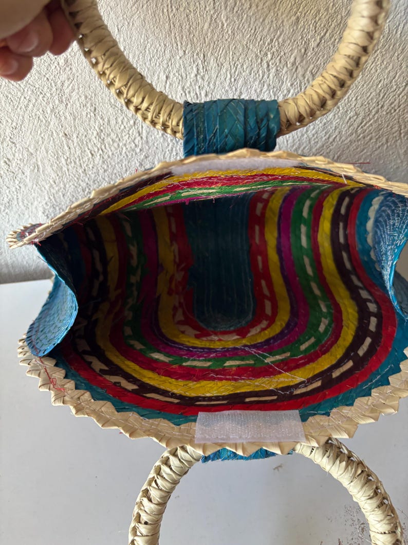 May include: A woven straw bag with a blue, yellow, red, green, and purple striped interior. The bag has two large, circular handles.