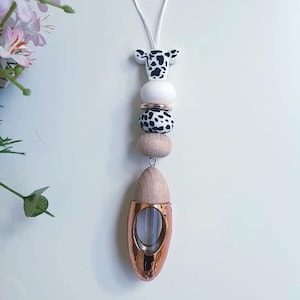May include: A car air freshener with a cow-shaped top, black and white beads, and a rose gold-coloured diffuser. The air freshener hangs from a white cord. The design includes wooden and white beads.