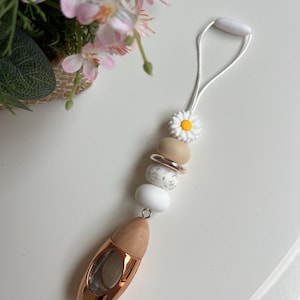 May include: An air freshener with a wooden top and a rose gold-coloured bottom. The air freshener is attached to a string with decorative beads, including a daisy, and a white loop. The background includes pink flowers.