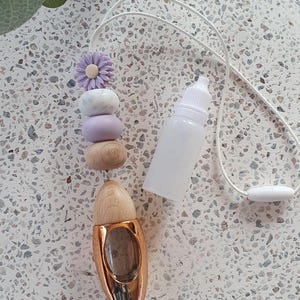 May include: A car air freshener featuring a gold-coloured shoe-shaped diffuser and a string of decorative beads in shades of purple, white, and wood. A small white bottle is also present. The air freshener is designed to hang in a vehicle.